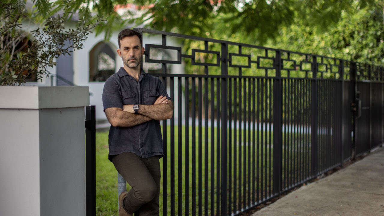 a man stands with folded arms leaning on a black iron fence