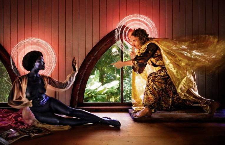 Photo of an editorial, stylized shoot featuring a nude woman in a robe and afro with a halo around her head. She's reaching out to a man in a silk patterned robe and cape