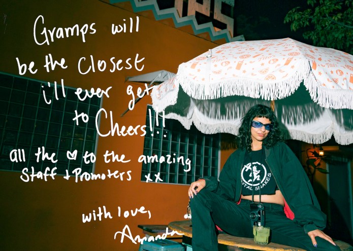 Photo of a woman sitting under an outdoor restaurant umbrella in front of an orange building. Text written on the photo reads, "Gramps will be the closest I'll ever get to Cheers! All the [heart symbol] to the amazing staff + promoters xx. With Love, Amanda"
