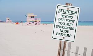 photo of a metal sign with green lettering reading, "Attention. Beyond this point you may encounter nude bathers." Behind the sign are the shores of a beach with a pastel-pink lifeguard tower in the distance