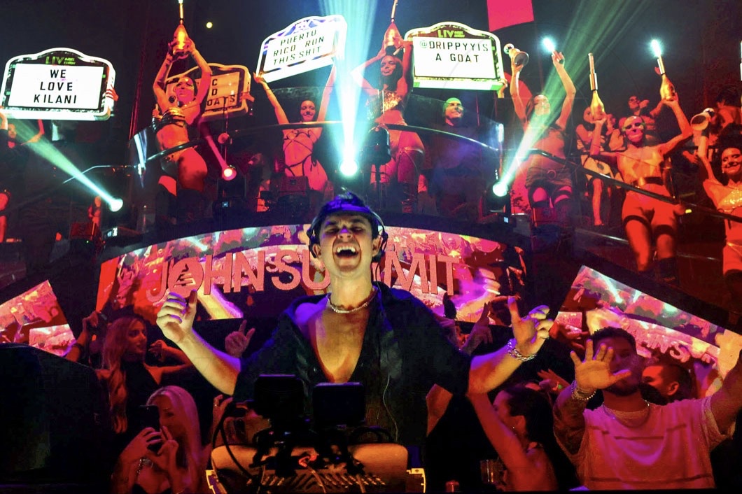 photo of DJ John Summit performing at a club with bottle girls holding up signs behind him