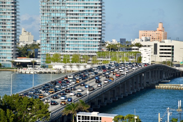 three lanes of cars sit in standstill traffic on a bridge over blue water, with high-rise towers in the background