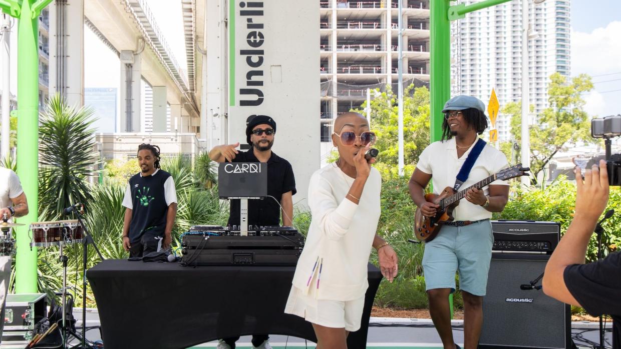 Photo of a bald woman on stage holding a microphone at an outdoors venue.