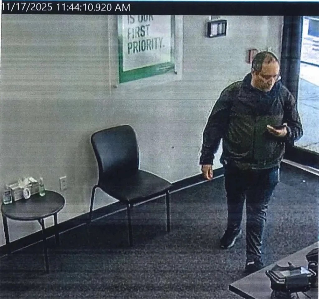 security still of a man with close-cropped hair and glasses, wearing long pants, a windbreaker, and sneakers, holding a cell phone