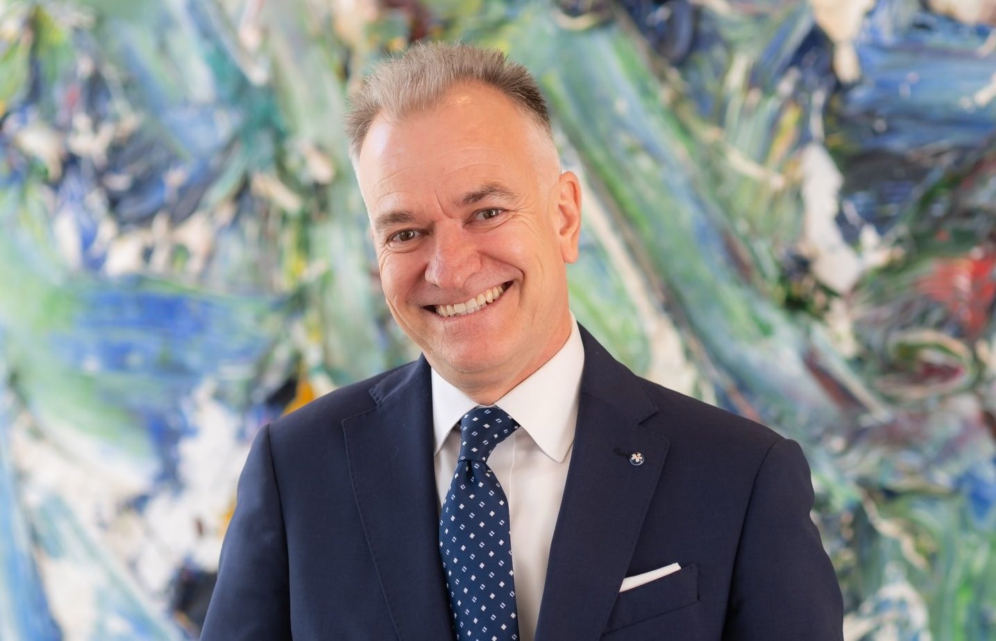 Horizontal image of a well-dressed man smiling graciously, standing in front of an abstract painting