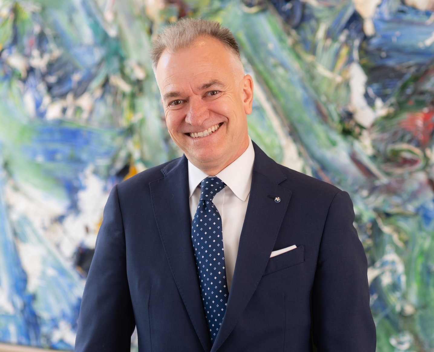 Horizontal image of a well-dressed man smiling graciously, standing in front of an abstract painting