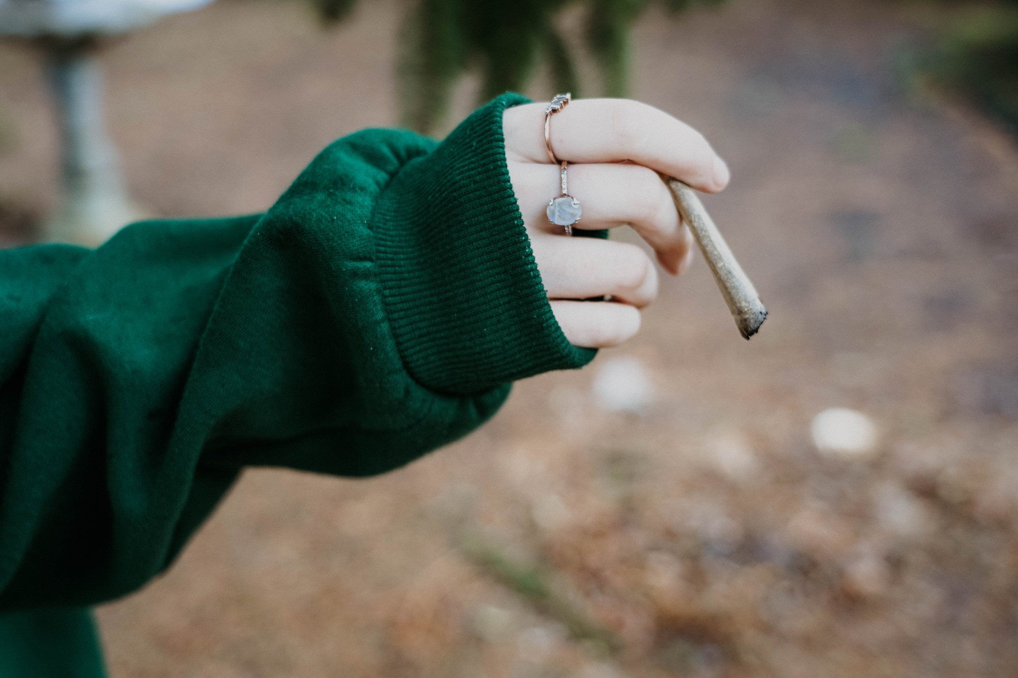 close-up of a pale female hand with several rings, emerging from a dark green sweatshirt sleeve,holding a burning marijuana joint