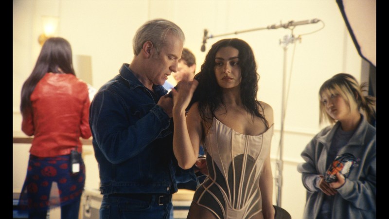film still from the movie The Moment. the image shows a man fixing Charli XCX's hair backstage at what appears to be a photoshoot