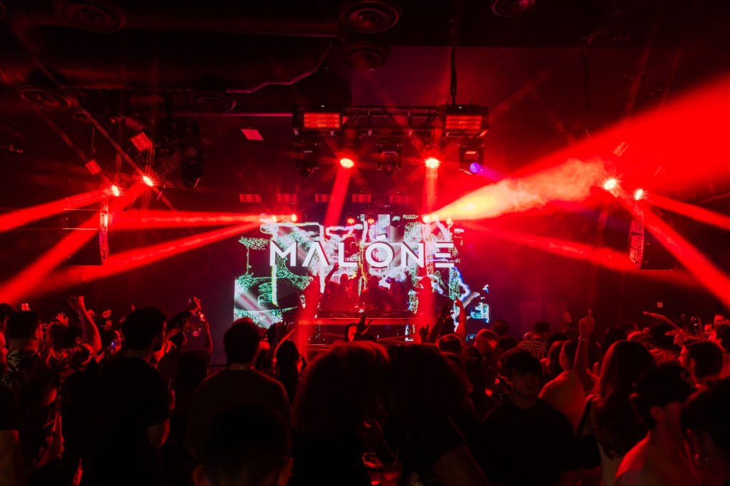 Photo of a venue with an LED light behind the stage that reads Malone. 