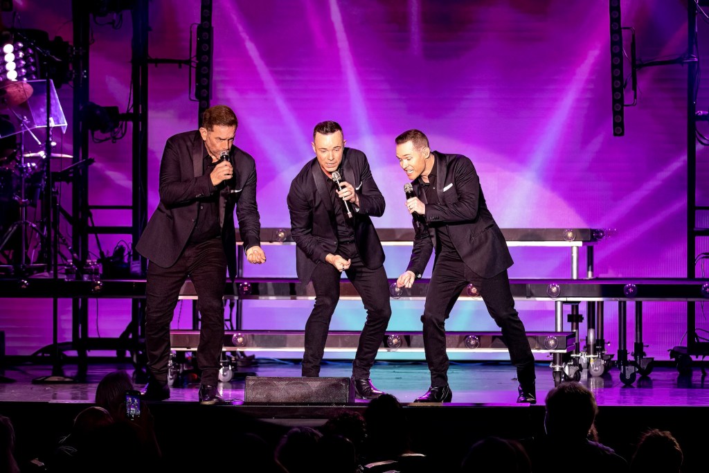 photo of three singers in suits performing on a stage with a purple background