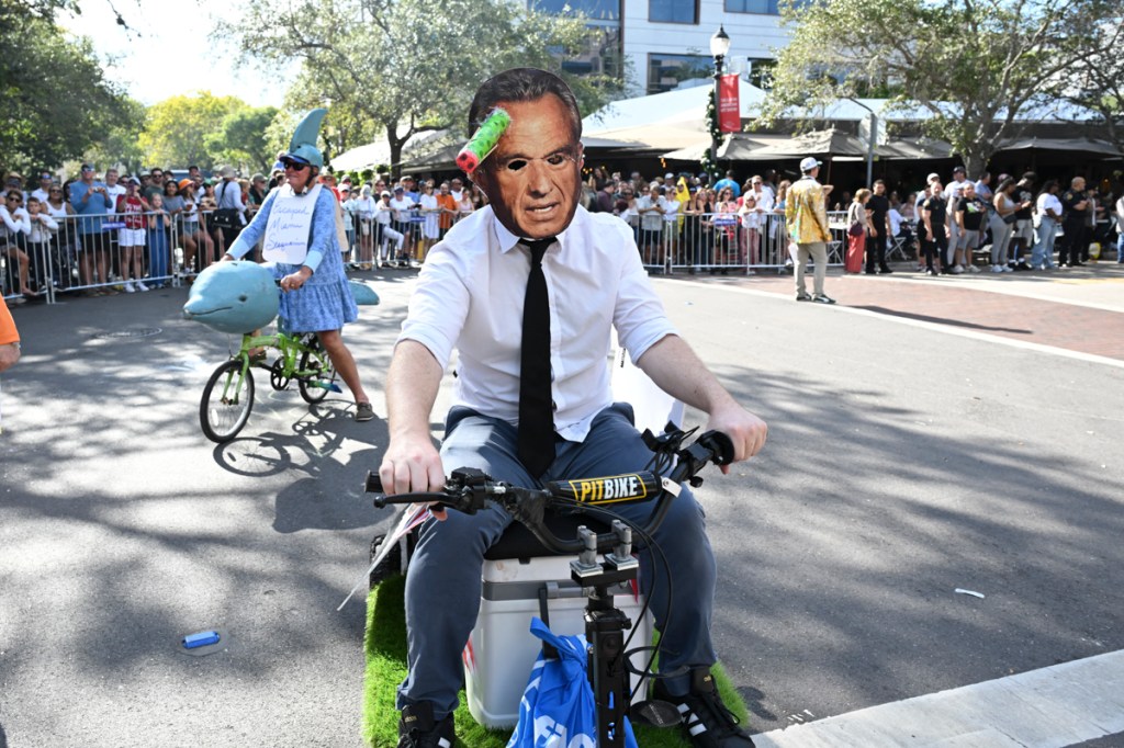 photo of a man riding a bicycle at a parade wearing a mask depicting Robert F. Kennedy, Jr.