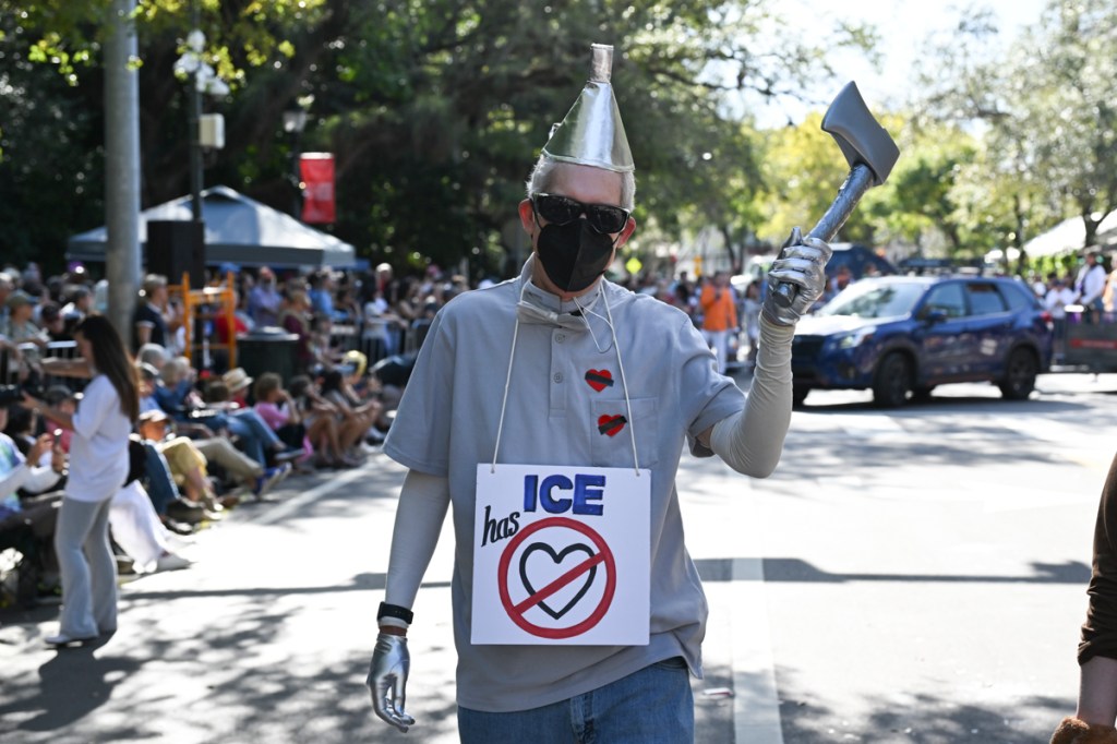 photo of a man dressed as the Tin Man in a parade wearing a sign reading "ICE has no [crossed over heart]"