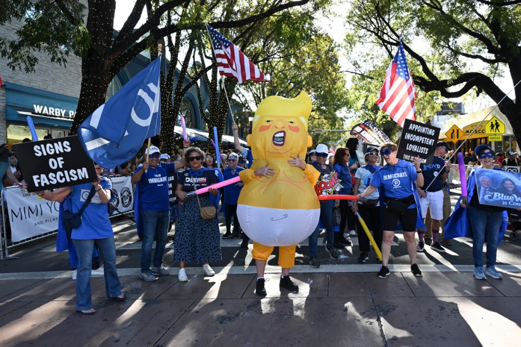 Photo of people on a parade route. The person in the center wears a costume of Donald Trump as a baby