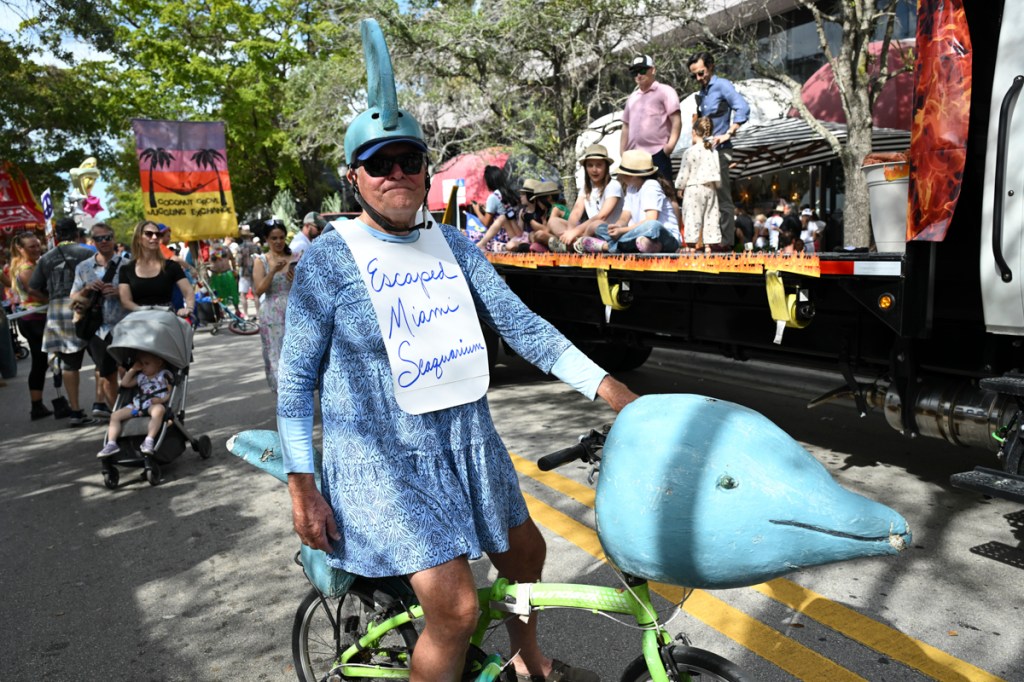 photo of a man at a parade riding a bike with a dolphin face on it. the man wears a sign reading, "escaped Miami seaquarium"