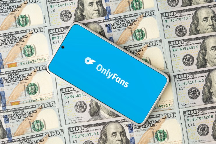 Photo of a smartphone on top of stacks of hundred-dollar bills. The phone displays the OnlyFans logo