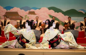 film still showing two geishas on a stage in front of men in uniforms