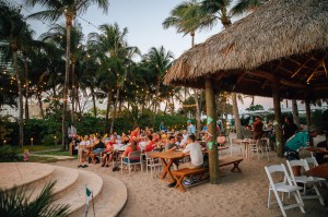 photo of a crowd sitting an picnic tables next to a tiki bar on sand