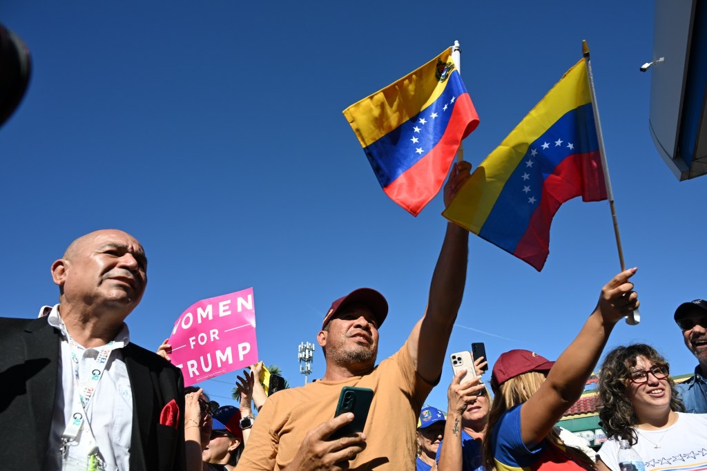 Photo of people at a rally, waving Venezuelan flags. A person in the back holds up a pink sign reading, "Women for Trump"