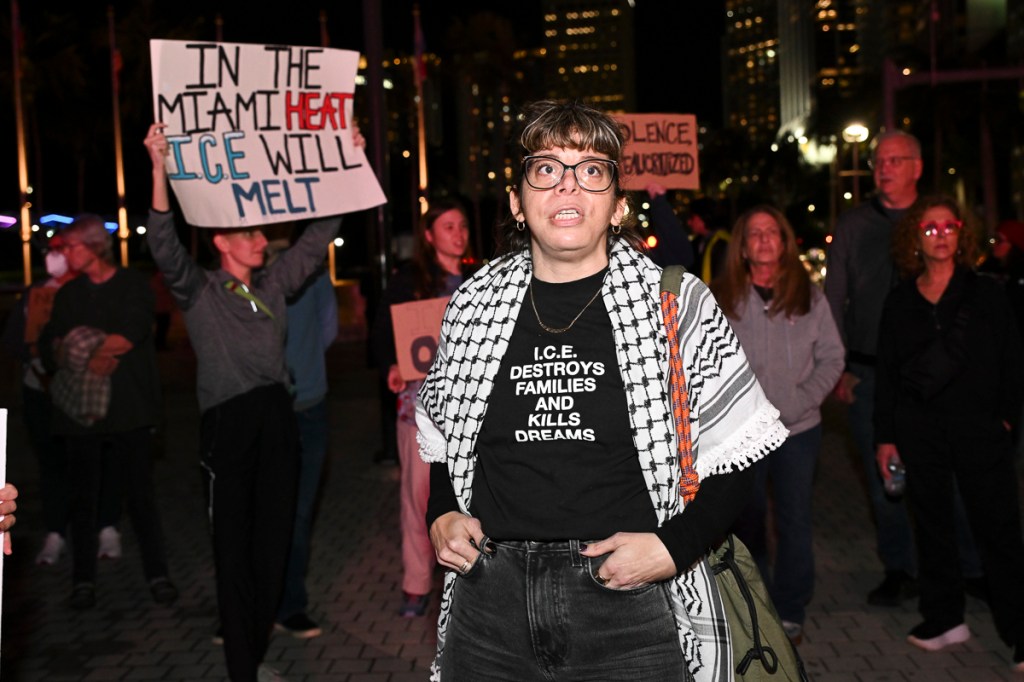 photo of protesters at a rally. a woman in the front wears a short reading, "ICE destroys families and kills dreams"