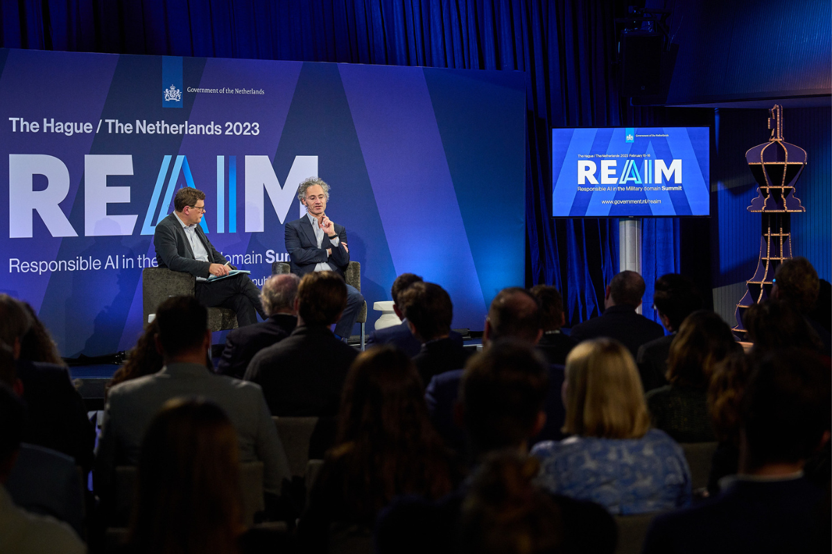 two men speak on a stage at a conference with a blue screen in the background