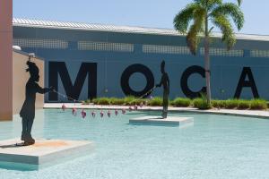 Photo of the facade of a museum with bold letters reading "MOCA." A pool is in the foreground with two black sculptures of children holding a string of fish