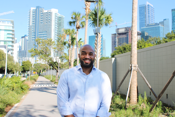 photo of a bearded Black man in a button-down shirt posing in a pedestrian pathway in with skyscrapers in the distance