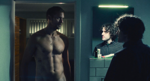 Film still of two men in a bathroom. The man on the left stands in a robe his bare chest and the man on the right looks at him as we see his reflection in a mirror