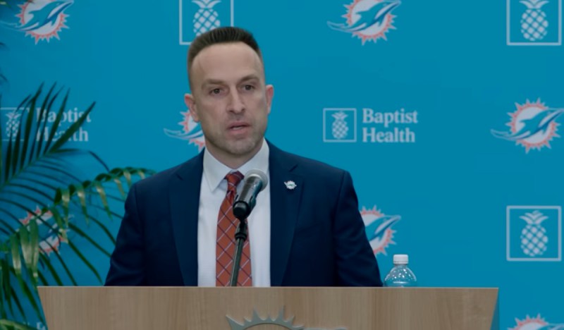 Jeff Hafley speaks at a podium at his introducery press conference. He is wearing a tie and jacket in front of a backdrop with Baptist Health and Miami dolphins logo