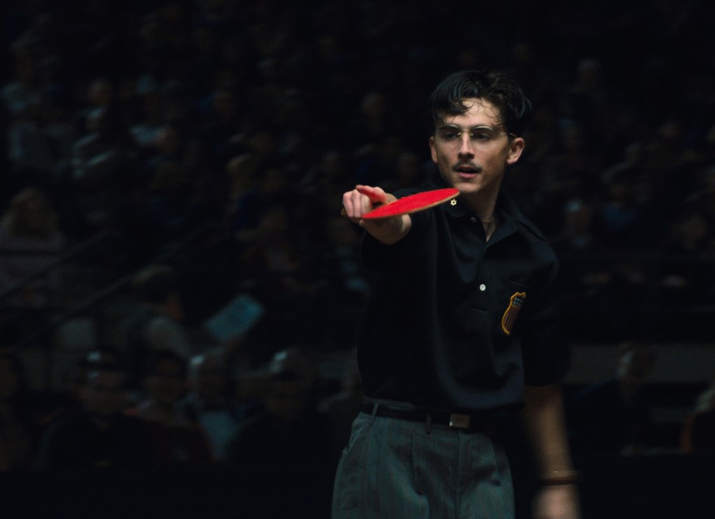 photo of actor Timothee Chalamet holding a ping pong paddle in front of a crowd in shadow