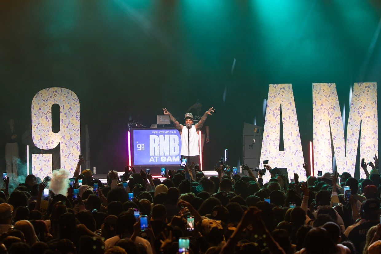 Photo of a man on stage in front of a crowd. The stage has letters that read 9am .