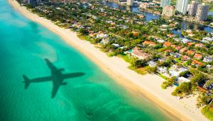 aerial photo of the shadow of a plane flying over the ocean in the direction of trees, houses, and skyscrapers in Miami
