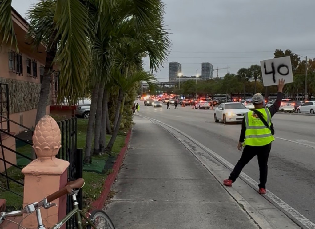 Photo of a man in a reflective vest waving down cars as he holds a sign reading, "40." The man's bike leans against a signpost nearby.