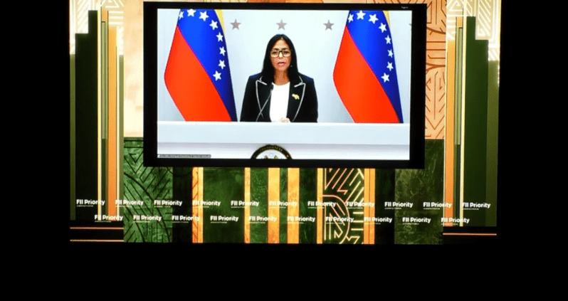 A photo of a bespectacled woman flanked by Venezuelan flags and appearing via video call on a large TV screen.