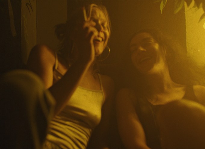 Still from the Erupcja showing two women in casual clothes laughing together as one smokes a cigarette. The film still is bathed in yellow light