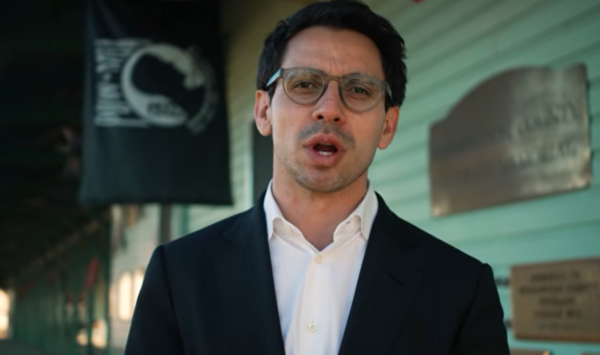 James Fishback wearing a white button down and blue sports jacket and glasses. He is speaking to the camera in an campaign video.