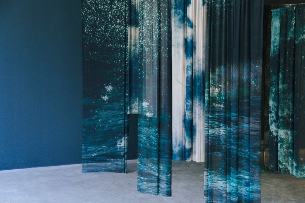 Photo of curtains hanging from the ceiling inside a gallery space in front of a blue wall. The curtains depict imagery of ocean water