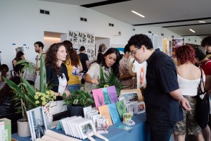Photo of a people walking around booths at a zine fair. In the center, two women at a booth show zines to a man looking at the options