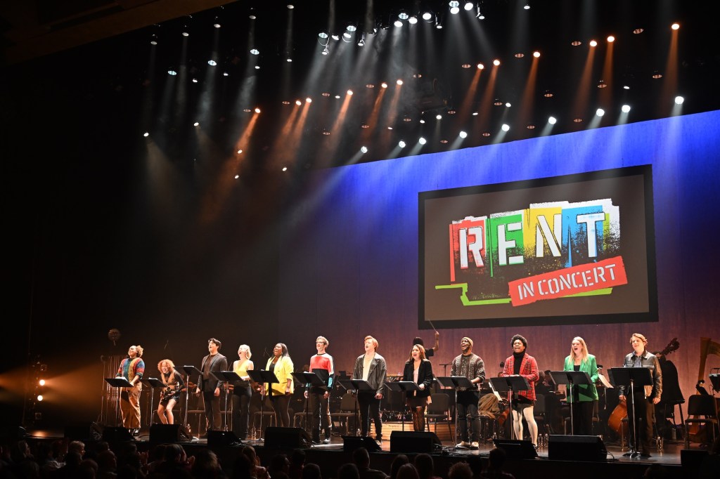 Photo of twelve singers standing in front of music stands on a stage during a concert. A screen behind them shows a projected logo reading, "RENT in concert"