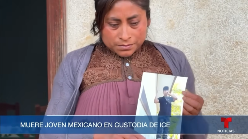 A screenshot of a Telemundo broadcast showing a woman solemnly holding a printed out photo of a child wearing a black Tshirt and pants.