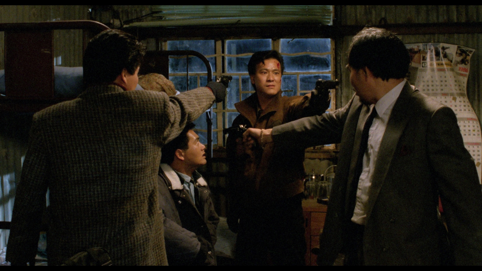 Still from the movie "City of Fire" showing a gun standoff between three men. One of the men points a gun at a man sitting in a chair in the middle of them