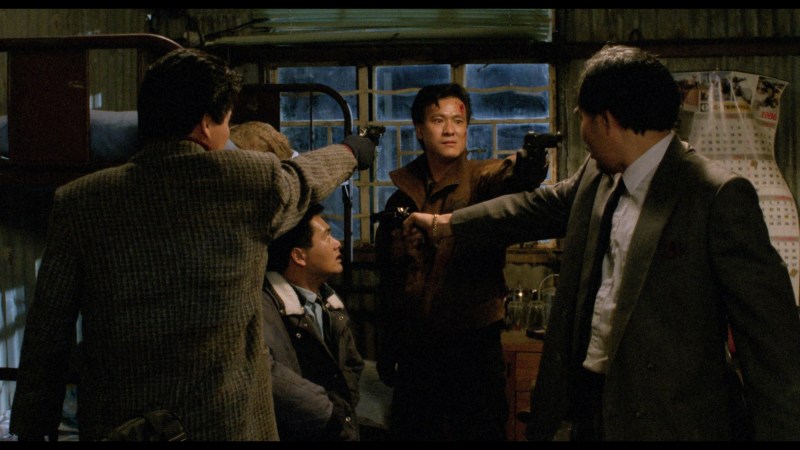 Still from the movie "City of Fire" showing a gun standoff between three men. One of the men points a gun at a man sitting in a chair in the middle of them