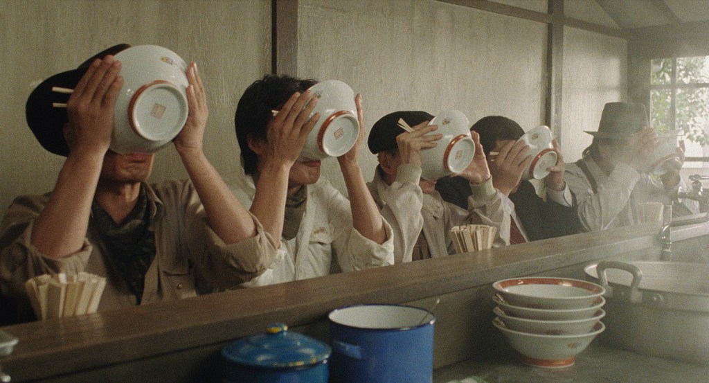 still from "Tampopo" film showing a line of people sitting at a lunch bar, with each drinking soup from bowls
