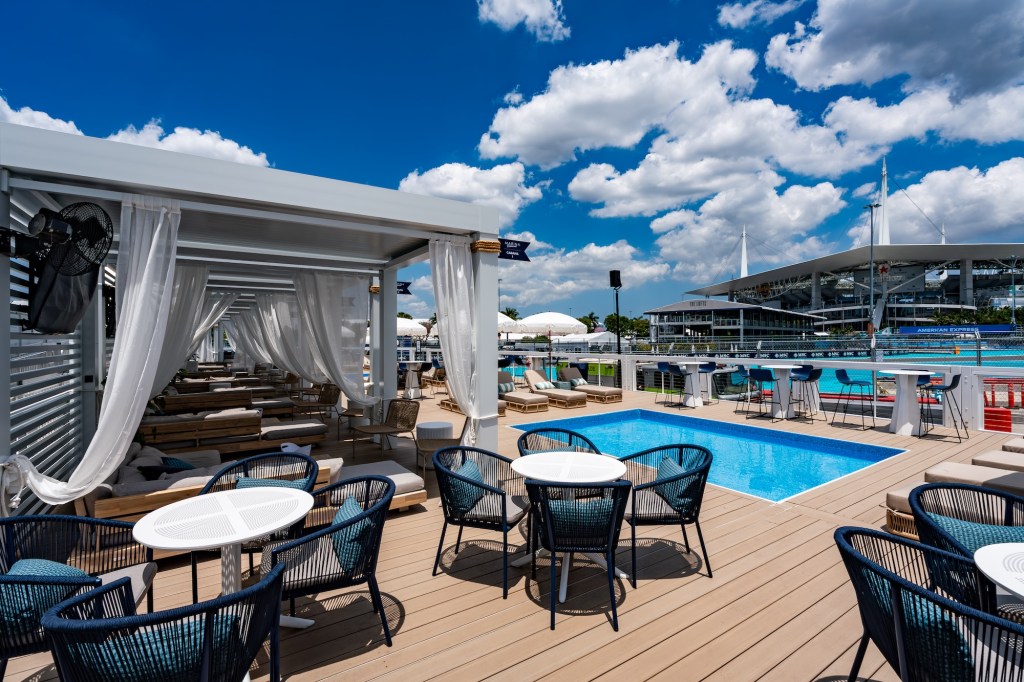 photo of a pool deck with cabanas, tables and chairs, and a stadium in the background