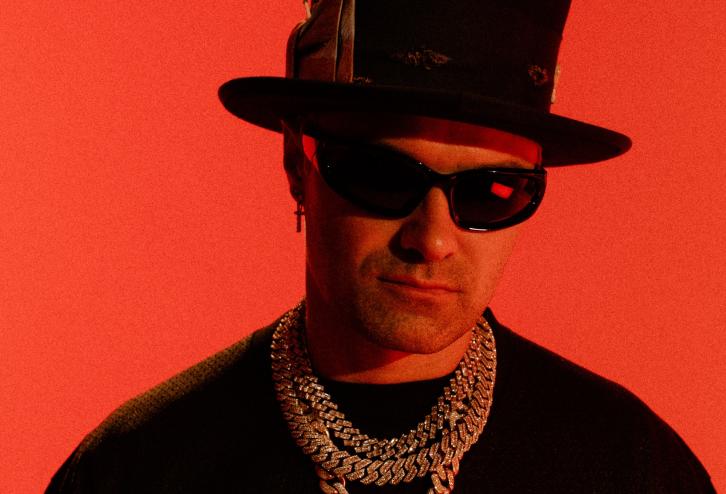 photo of artist Alec Monopoly posing in a top hat, thick necklaces, and shades against a bright-red backdrop