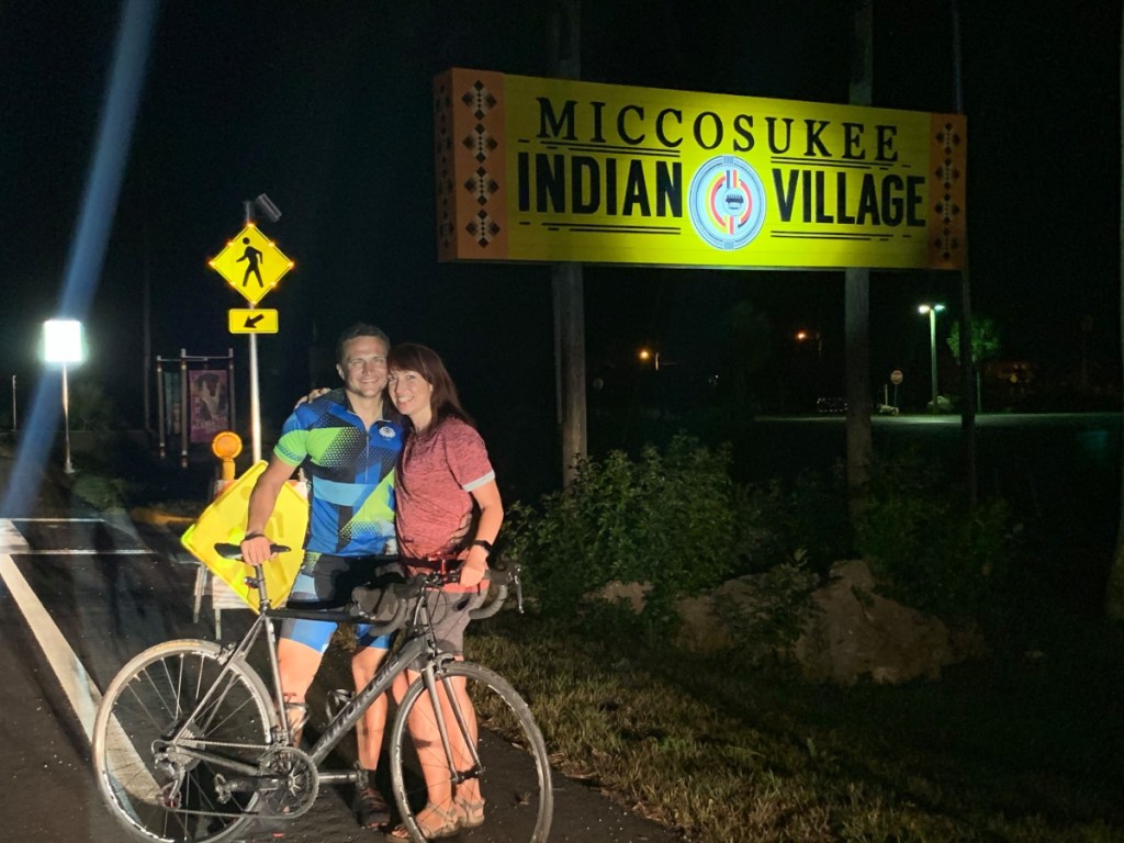 A photo of a man and woman with bikes next to a large sign that reads "Miccosukee Indian Village"