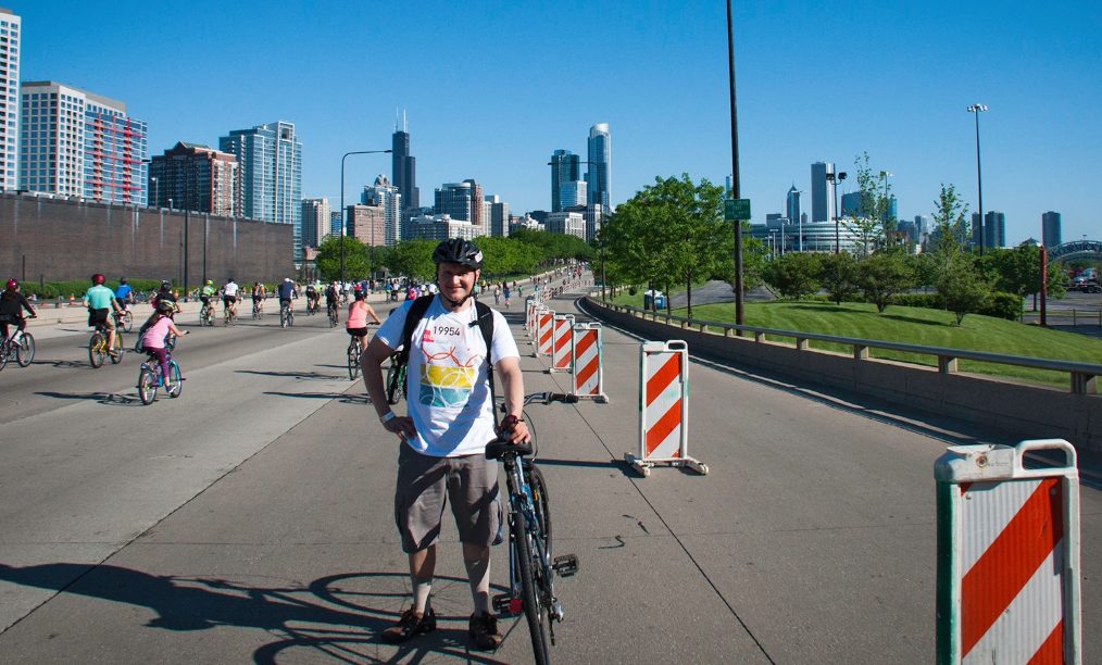 A photo of a man standing with his bike with the Chicago skyline in the background.