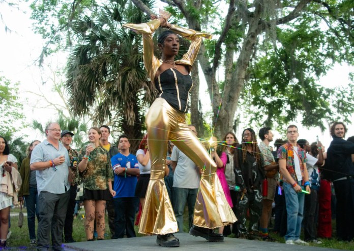 photo of a person in a black and gold bodysuit walking a makeshift runway in front of a crowd outside surrounded by trees