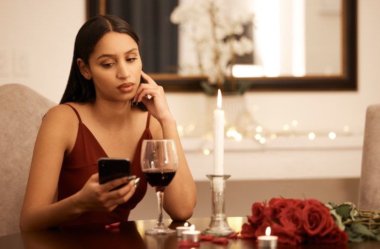 Photo of a woman in a slinky red dress staring at her phone with an unimpressed look on her face. In front of her is a table with