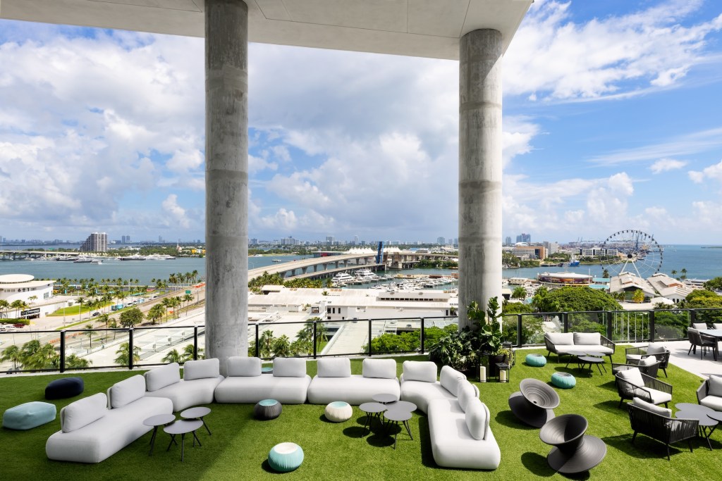 Photo of a hotel rooftop showing outdoor couches and lounge chairs beneath industrial concrete columns and ceiling. The Miami cityscape, a highway, and a ferris wheel can be seen in the background