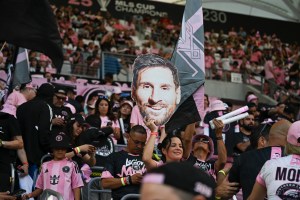 photo of fans holding up a cardboard cutout of Lionel Messi's face in the stands at an Inter Miami FC match